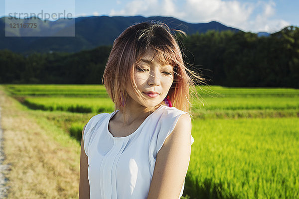 Eine junge Frau in einem weißen Hemd  mit vom Wind verwehten Haaren  steht im Freien an Reisfeldern.