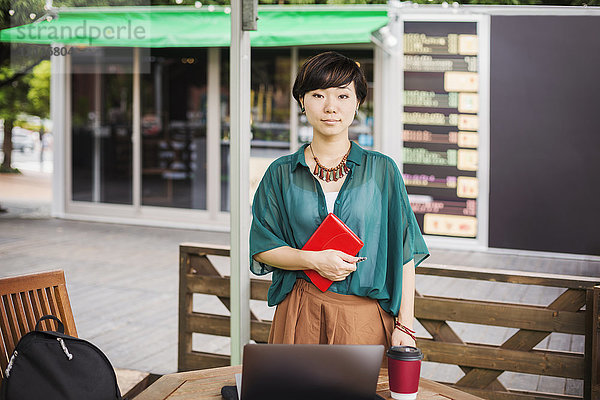 Frau mit schwarzen Haaren und grünem Hemd steht draußen in einem Straßencafé und schaut in die Kamera.