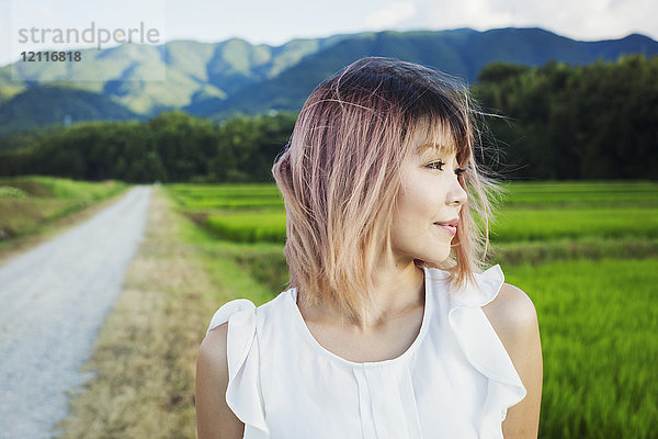 Eine junge Frau steht im Freien an Reisfeldern mit grünen Trieben und einer Berglandschaft.