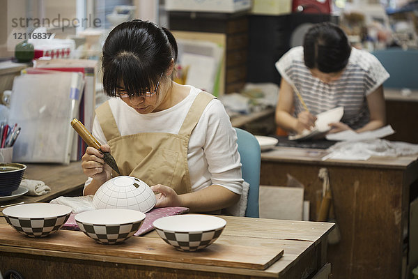 Frau  die in einer japanischen Porzellanwerkstatt arbeitet und mit dem Pinsel geometrische Muster auf weiße Schalen malt.