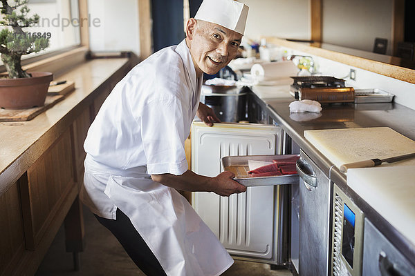 Chefkoch  der in einem japanischen Sushi-Restaurant an der Theke arbeitet und ein Metalltablett mit Fisch in den Kühlschrank stellt.