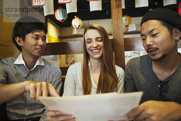Drei lächelnde Menschen  eine Frau und zwei Männer  sitzen Seite an Seite an einem Tisch in einem Restaurant und schauen auf die Speisekarte.
