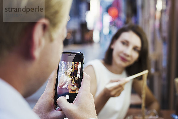 Mann fotografiert mit seinem Smartphone eine Frau  die an einem Tisch in einem asiatischen Restaurant sitzt  Essstäbchen benutzt und Nudeln isst.