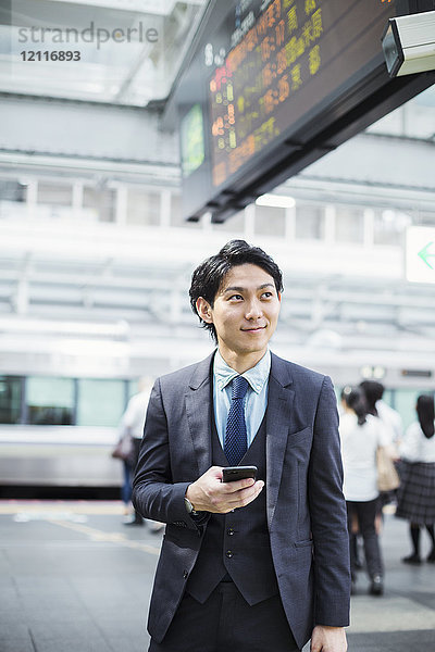 Geschäftsmann im Anzug  steht auf dem Bahnsteig und hält ein Mobiltelefon in der Hand.