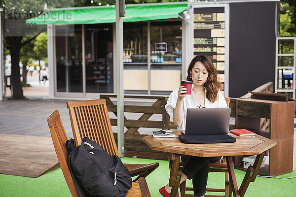 Frau mit schwarzen Haaren und weißem Hemd  die in einem Straßencafé vor einem Laptop am Tisch sitzt und arbeitet.