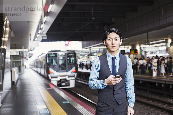 Geschäftsmann in blauem Hemd und Weste steht auf dem Bahnsteig des Bahnhofs  hält Mobiltelefon in der Hand und schaut in die Kamera.