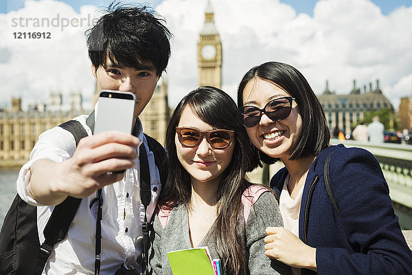 Lächelnder Mann und zwei Frauen mit schwarzen Haaren  die sich mit einem Smartphone selbstständig machen  stehen auf der Westminster Bridge über die Themse  London  mit dem Houses of Parliament und Big Ben im Hintergrund.