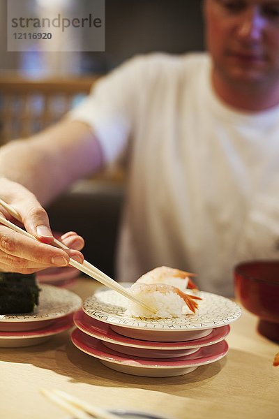Nahaufnahme eines Mannes  der in einem asiatischen Restaurant am Tisch sitzt und mit Stäbchen ein Stück Sushi von einem Teller aufhebt.