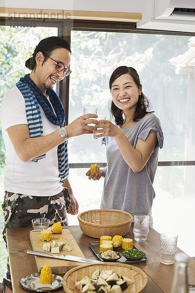 Lächelnder Mann und lächelnde Frau stehen drinnen an einem mit Essen gedeckten Tisch  halten Trinkgläser in der Hand und stoßen an.