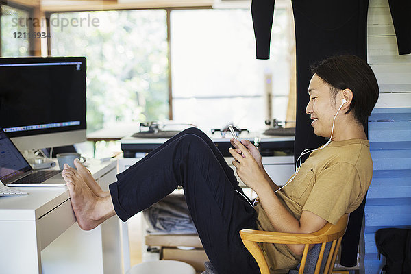 Seitenansicht eines Mannes  der drinnen an einem Schreibtisch mit Computer sitzt  Beine hochgelagert  barfuß  Handy in der Hand  Kopfhörer tragend.