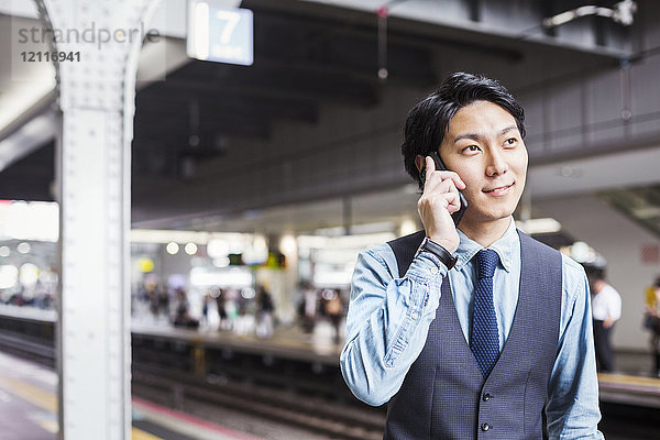 Geschäftsmann in blauem Hemd und Weste steht auf dem Bahnsteig des Bahnhofs und telefoniert mit einem Mobiltelefon.