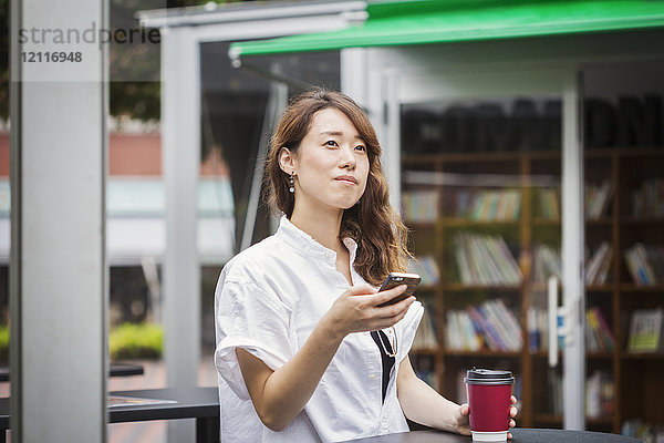 Frau mit braunen Haaren  die ein weißes Hemd trägt  im Freien steht und Handy und Pappbecher hält.