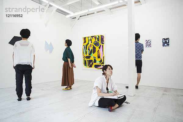 Frau mit schwarzen Haaren sitzt in der Kunstgalerie mit Stift und Papier auf dem Boden und betrachtet moderne Malerei  drei Personen stehen vor Kunstwerken.