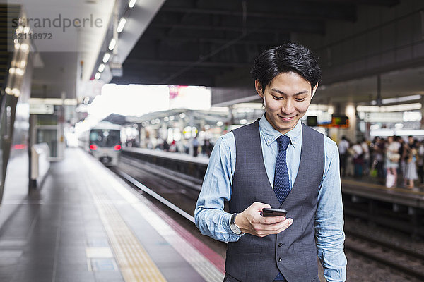 Geschäftsmann in blauem Hemd und Weste steht auf dem Bahnsteig des Bahnhofs und hält ein Mobiltelefon in der Hand.