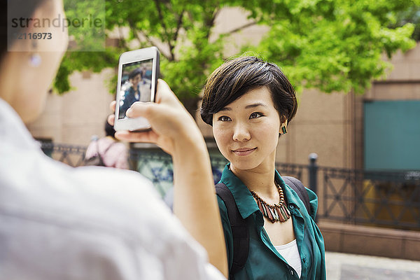 Zwei Frauen mit schwarzen Haaren in weißem und grünem Hemd stehen im Freien  fotografieren mit dem Handy und lächeln.