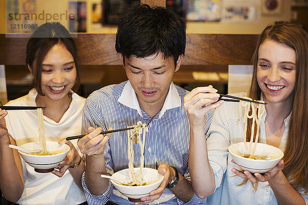 Drei lächelnde Menschen sitzen nebeneinander an einem Tisch in einem Restaurant und essen mit Stäbchen aus Schalen.