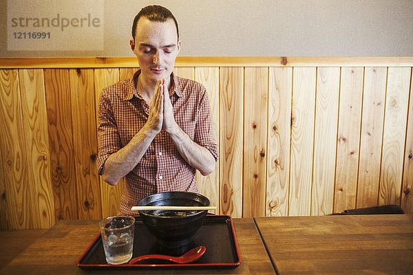 Ein westlicher Mann in einem Nudelrestaurant  die Hände im Gebet und mit geschlossenen Augen  der vor seiner Mahlzeit ein traditionelles Itadakimasu spricht.