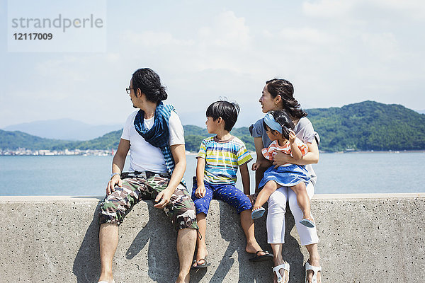Eine Familie  ein Mann  ein Junge und eine Frau mit einem jungen Mädchen auf dem Schoß  die Seite an Seite auf einer Mauer am Meer sitzen.