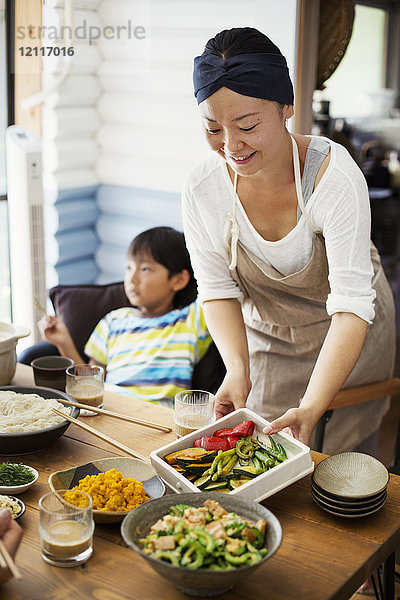 Lächelnde Frau mit Schürze  die Schüsseln mit Salat und Gemüse auf den Tisch stellt  im Hintergrund sitzt ein Junge.