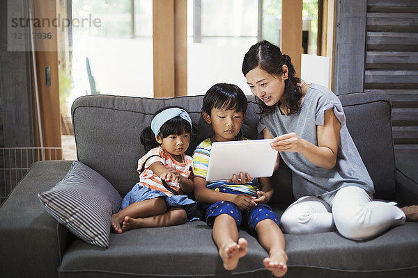 Frau  Junge und junges Mädchen sitzen auf einem grauen Sofa und schauen auf ein digitales Tablett.