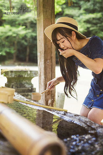 Junge Frau in blauem Kleid und Hut mit Bambuswasser-Handwaschbecken im Shinto Sakurai-Schrein  Fukuoka  Japan.