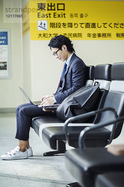 Geschäftsmann mit Anzug und Brille  der am Bahnhof sitzt und am Laptop arbeitet.