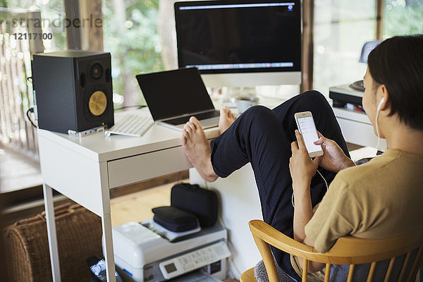 Mann sitzt drinnen an einem Schreibtisch mit Computer  Beine hochgelagert  barfuß  Handy in der Hand.