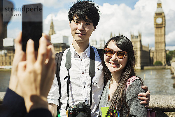 Lächelnde Frau mit schwarzen Haaren  die ein Paar mit einem Smartphone fotografiert  das auf der Westminster Bridge über die Themse in London steht  mit dem Houses of Parliament und Big Ben im Hintergrund.