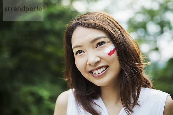 Porträt einer jungen Frau mit braunen Haaren  auf die Wange gemalte japanische Flagge  die in die Kamera lächelt.