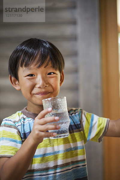 Porträt eines Jungen mit schwarzen Haaren  der ein gestreiftes T-Shirt trägt  ein Trinkglas hält und in die Kamera lächelt.