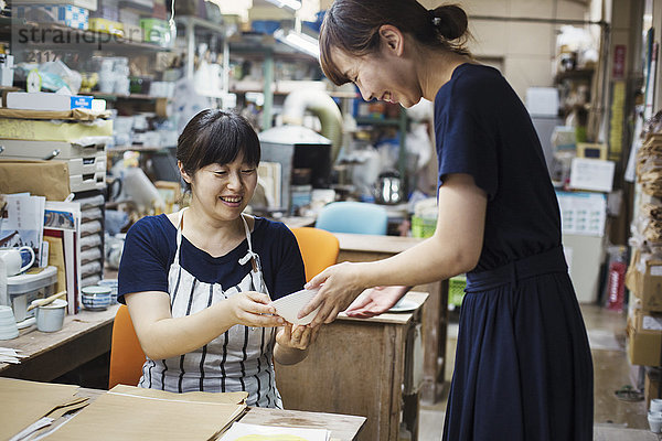 Zwei lächelnde Frauen sitzen und stehen in einer Werkstatt und schauen auf eine japanische Porzellanschüssel.