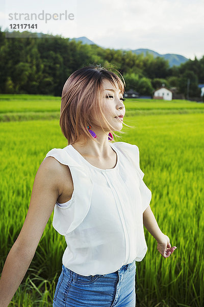 Eine junge Frau in weißem Hemd und Jeans mit ausgestreckten Händen steht auf freiem Feld an Reisfeldern.