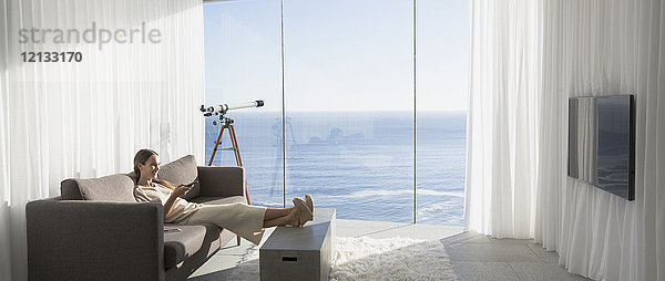 Frau entspannt sich mit hochgelegten Füßen vor dem Fernseher in einem modernen  luxuriösen Wohnzimmer mit sonnigem Meerblick