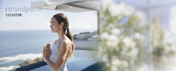 Gelassene brünette Frau  die Yoga praktiziert und mit den Händen in der Herzmitte auf einer sonnigen Terrasse mit Blick auf das Meer meditiert