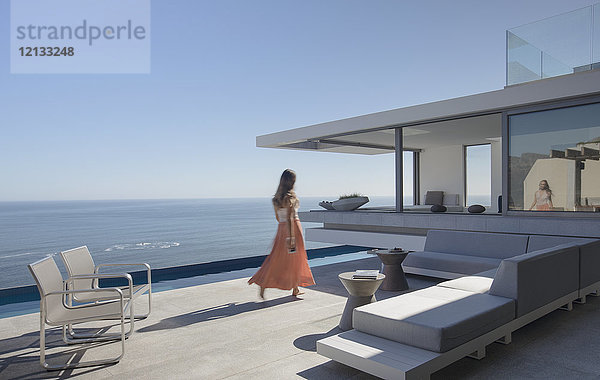 Frau im Kleid  die auf einer sonnigen  modernen  luxuriösen Wohnung spazieren geht  die eine Außenterrasse mit Meerblick hat