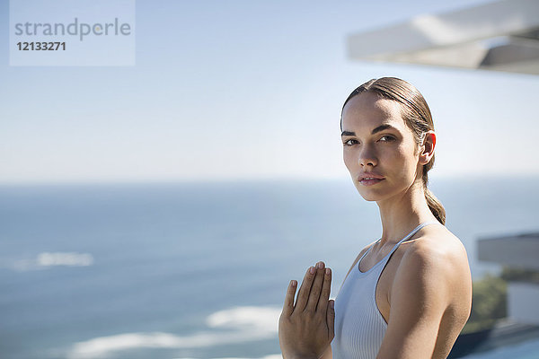 Porträt heitere Frau übt Yoga mit Händen in der Mitte des Herzens auf sonnigen Terrasse mit Blick auf das Meer