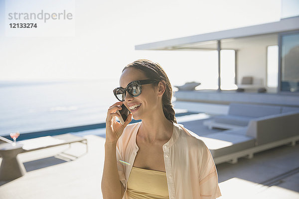 Lächelnde Frau  die auf einer sonnigen  modernen und luxuriösen Terrasse mit einem Smartphone telefoniert