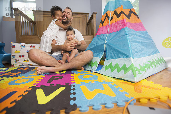 Vater sitzt auf dem Spielzimmerboden und hält Sohn und Tochter