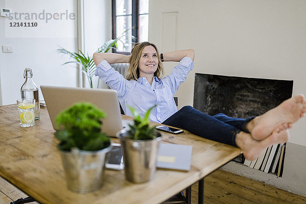 Lächelnde Frau sitzt zu Hause am Schreibtisch mit erhobenen Füßen