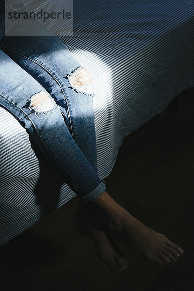 Nahaufnahme einer Frau  die zu Hause auf dem Bett sitzt und zerrissene Jeans trägt.