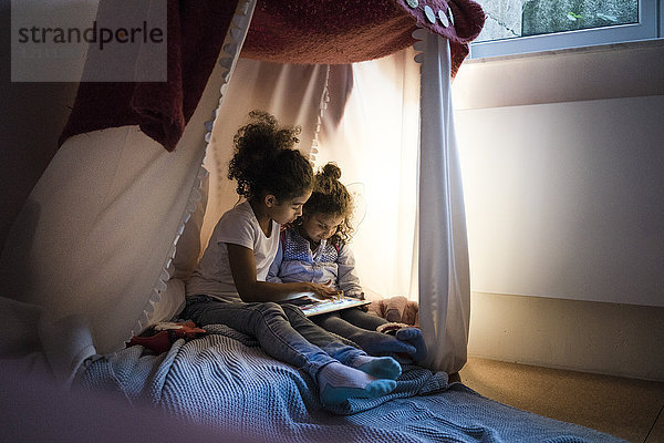 Zwei Schwestern sitzen im dunklen Kinderzimmer und schauen auf das digitale Tablett.
