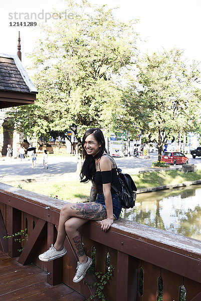 Porträt einer fröhlich tätowierten Frau auf einer Holzbrücke sitzend