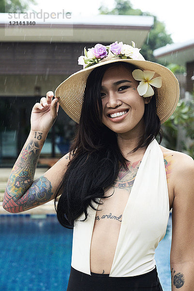 Porträt einer lächelnden Frau mit Blume im Haar  die einen Strohhut am Swimmingpool trägt.