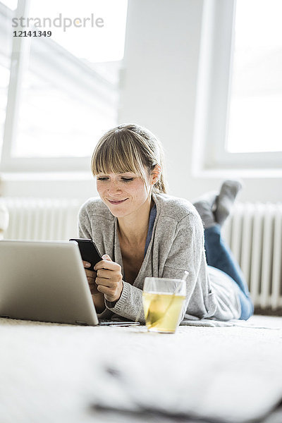 Lächelnde Frau auf dem Boden liegend mit Handy und Laptop