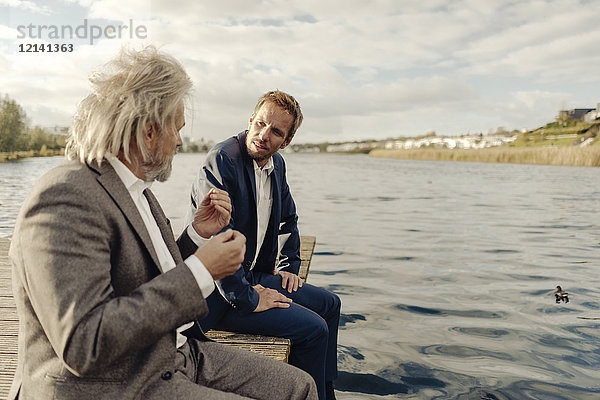 Zwei Geschäftsleute  die auf einem Steg am See sitzen und sich unterhalten.