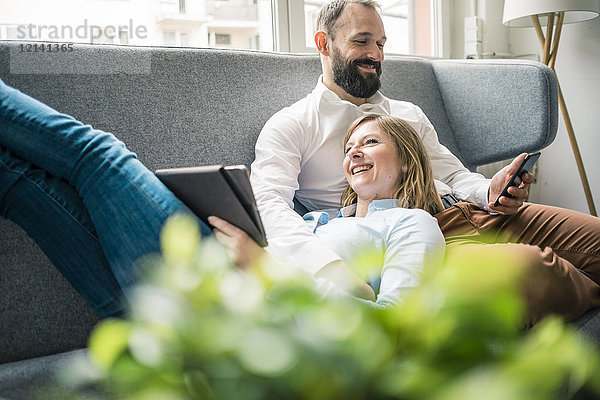 Lächelndes Paar mit Tablett und Handy auf der Couch