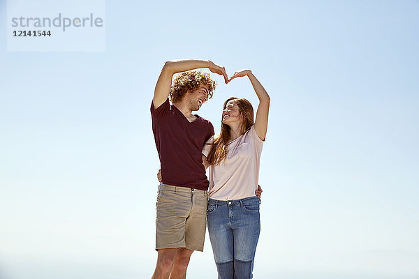 Glückliches junges Paar  das mit den Händen unter blauem Himmel ein Herz formt.