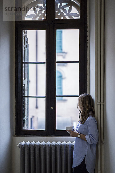 Frau mit Tasse Kaffee zu Hause aus dem Fenster schauend