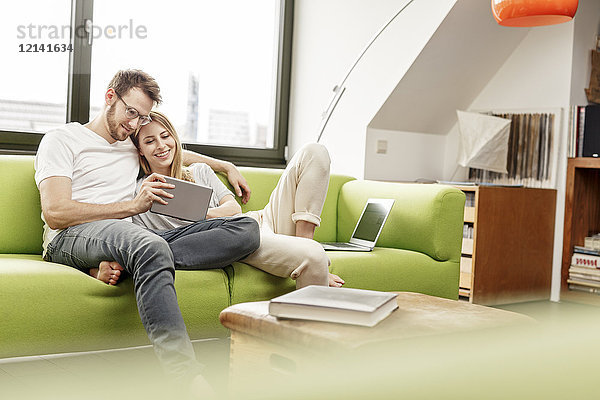 Lächelndes junges Paar auf der Couch im Wohnzimmer zu Hause  das sich die Tablette teilt.