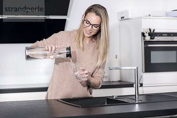 Lächelnde blonde Frau gießt Wasser in ein Glas in der Küche